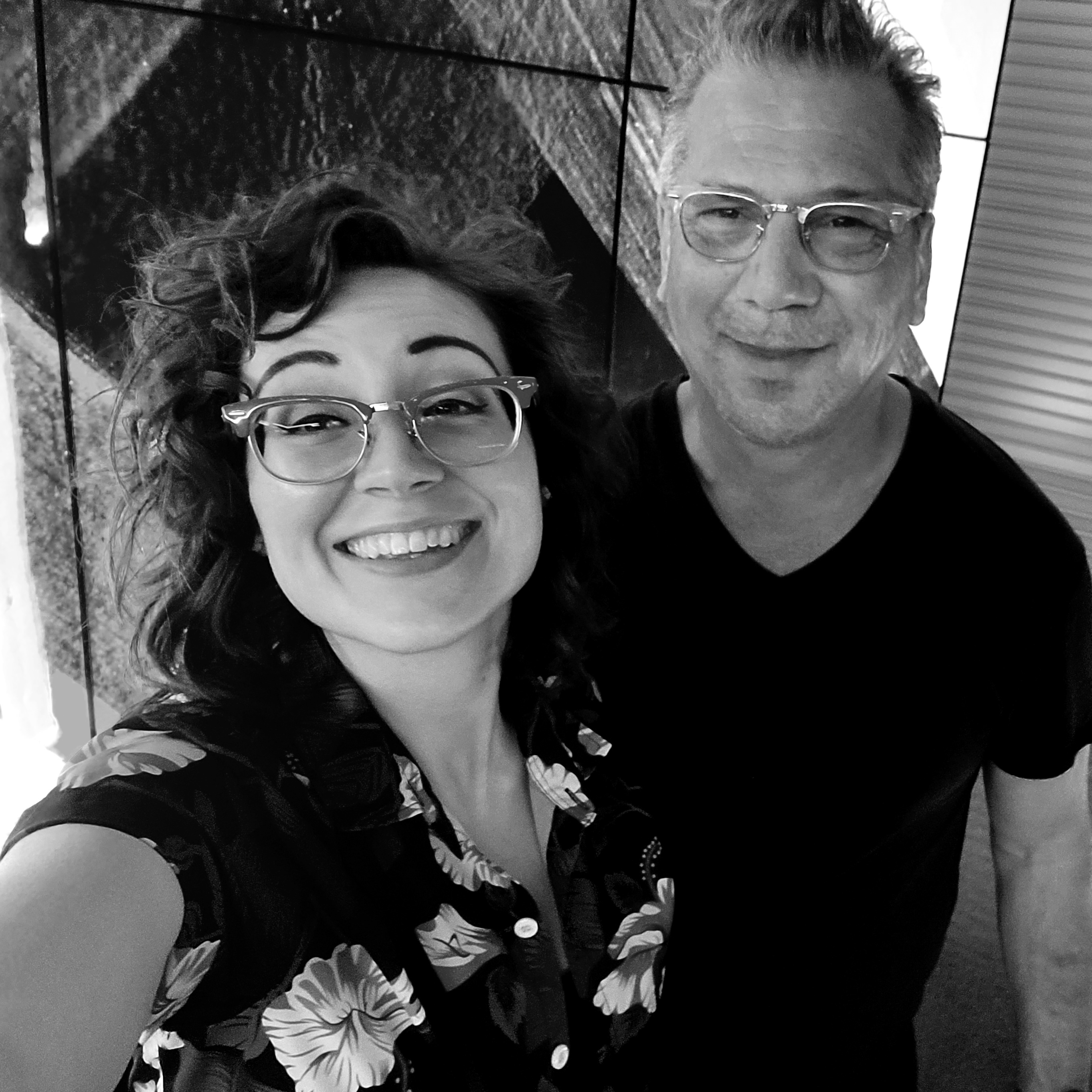 A selfie of Brenna and her husband David. Brenna is a white woman in her thirties with wavy hair and glasses. David is a white man in his fifties with short hair and glasses. They are smiling and standing in front of an LED wall.